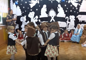 Na tle bożonarodzeniowej dekoracji w kółeczku tańczą dziewczynki oraz chłopcy w strojach ludowych.